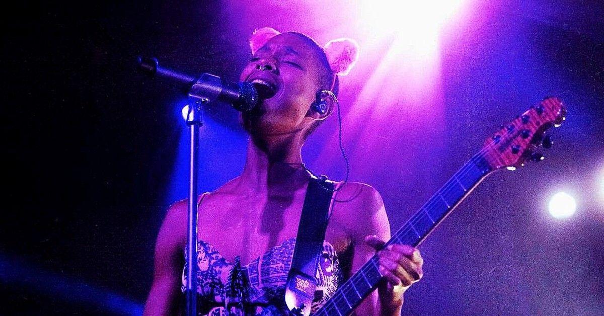 Willow Smith em luzes roxas no palco cantando enquanto toca guitarra